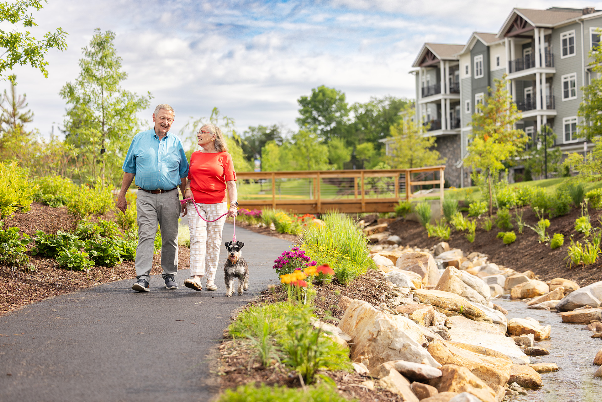 An elderly couple walks their dog on a paved path in a park-like setting.