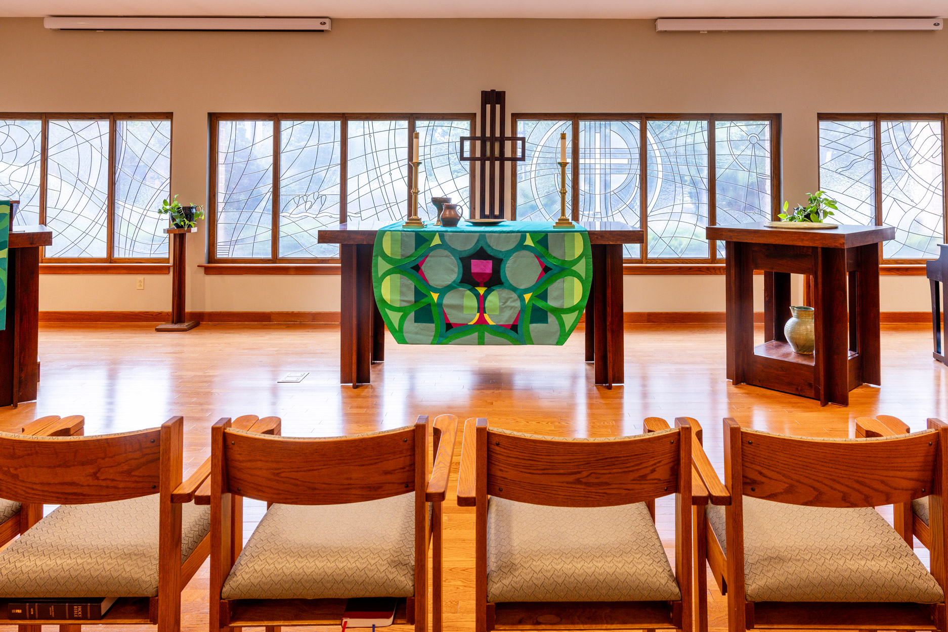 A chapel interior with a vibrant altar, wooden chairs, and large stained-glass windows.