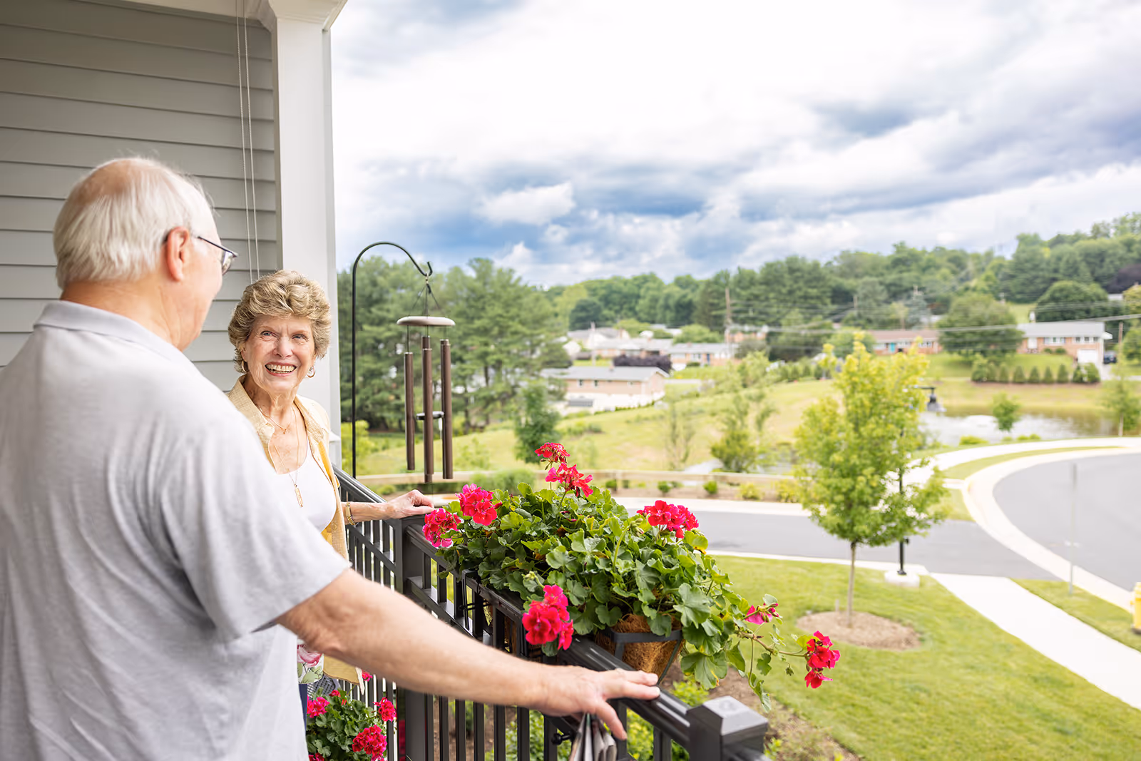 Elderly couple smiling and standing on a balcony with red flowers, overlooking a suburban neighborhood with greenery under a cloudy sky.
