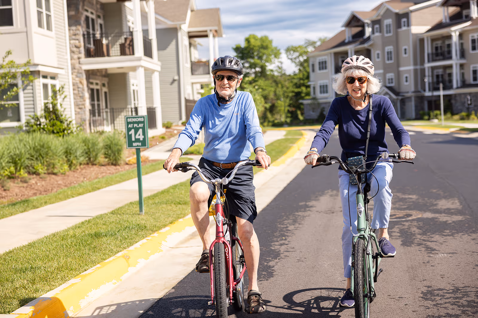 Senior man and woman wearing helmets and sunglasses riding bicycles on a residential street with apartment buildings in the background.