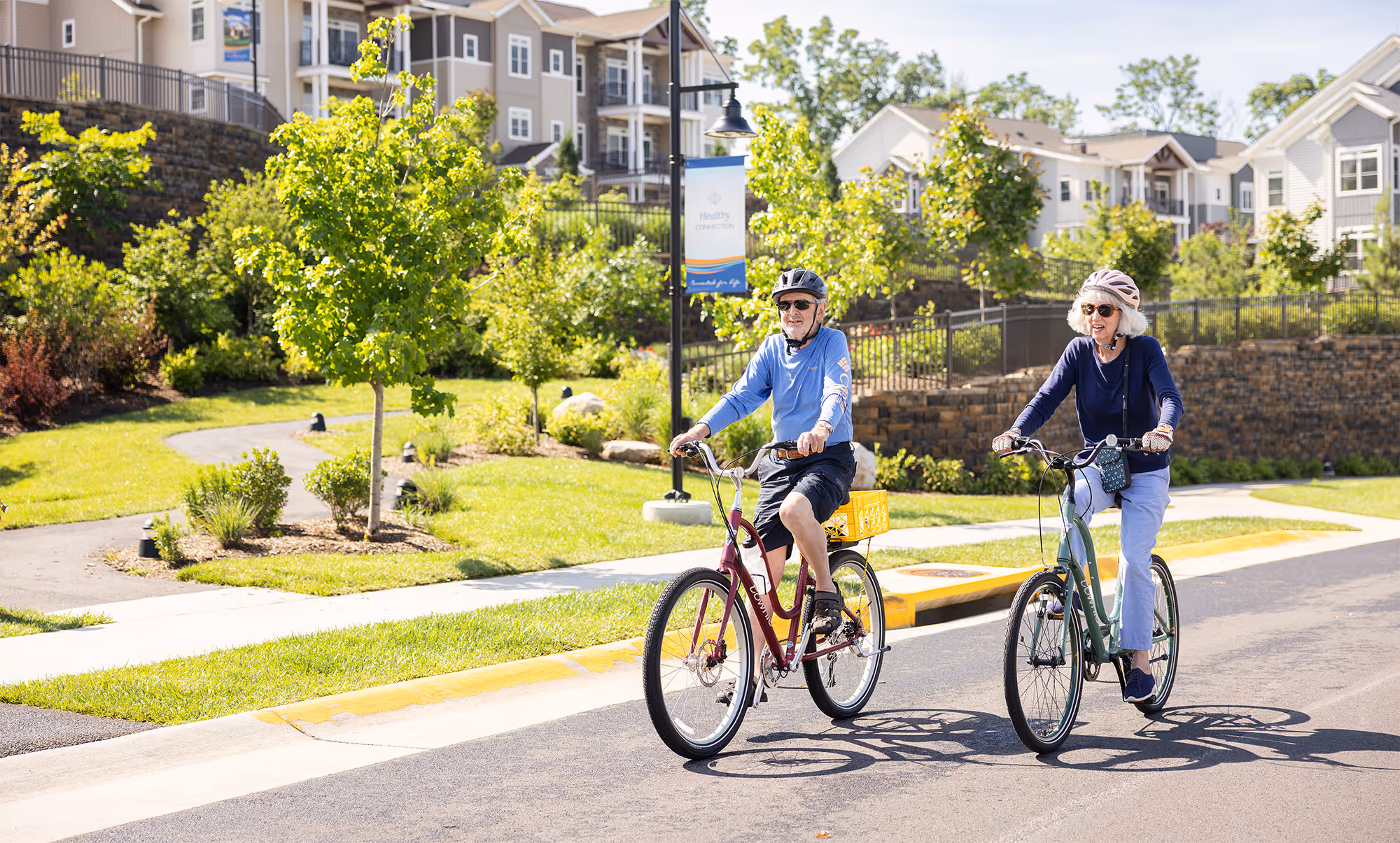 Senior man and woman wearing helmets cycling on a sunny residential street with greenery and houses in the background.