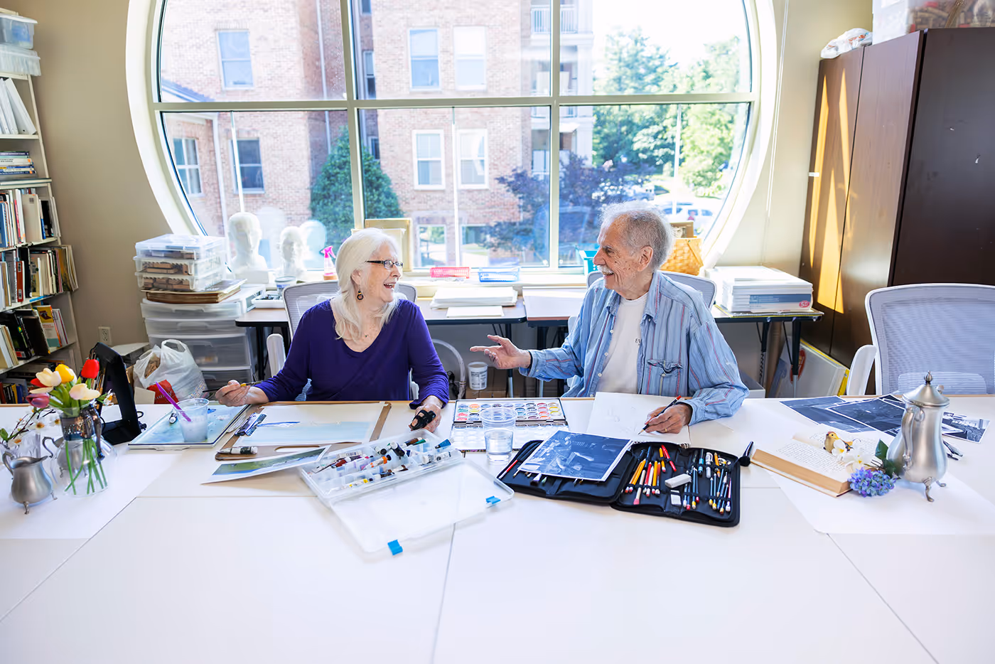 Two elderly people sitting at a table engaged in painting and drawing, smiling and talking in a bright room with a large round window.