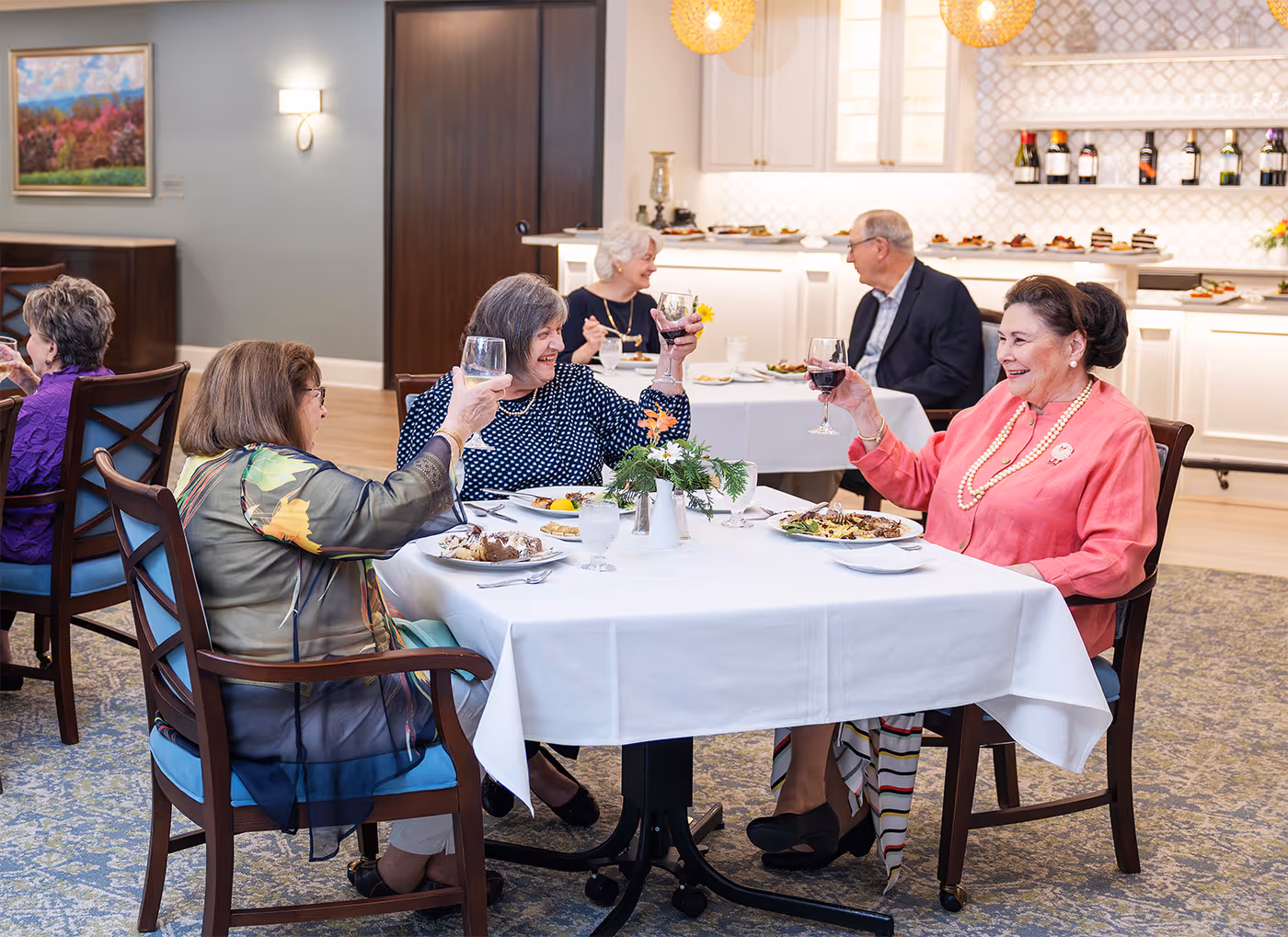 Three elderly women sitting at a dining table raising their wine glasses in a toast during a meal in a bright, elegant dining room.
