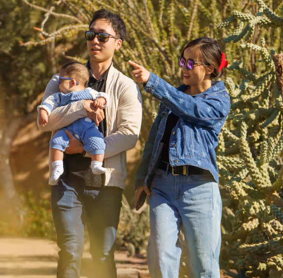 Two parents and their baby. One is pointing with a smile and the other is holding the baby. All of them have sunglasses on, even the baby. Fantastic vibes.