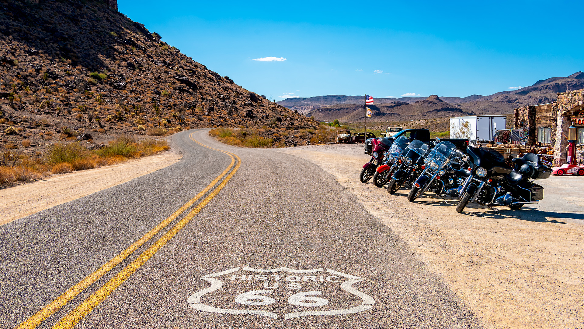 Motorcycles parked along Historic Route 66 in a desert landscape, with a winding road, American flag, rustic buildings, and mountains in the background under a clear blue sky.
