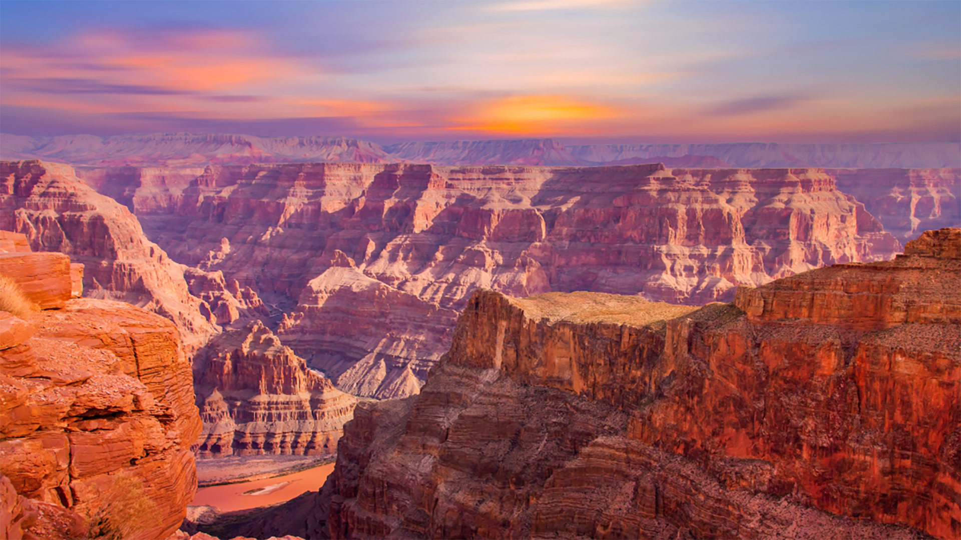Vibrant sunset over the Grand Canyon, casting warm hues across the layered red rock cliffs and winding Colorado River below.