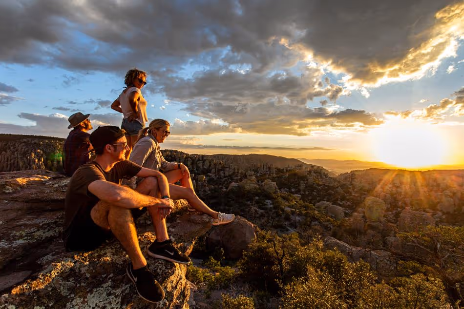 Four people sitting and standing on rocky cliffs watching a sunset over a forested valley with cloudy sky.