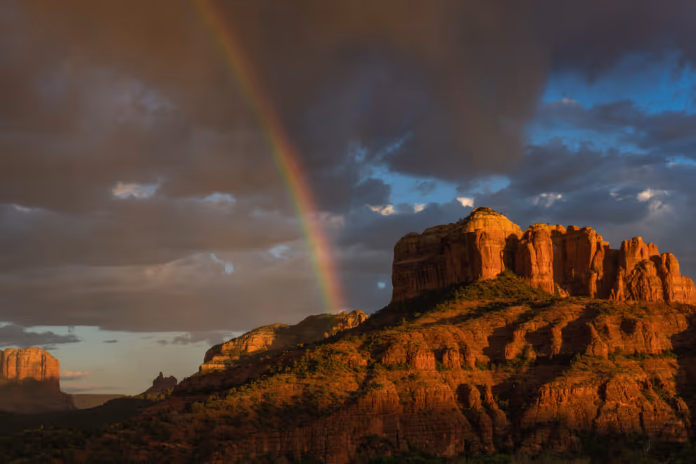 Rainbow arching over sunlit red rock formations under a dark cloudy sky.