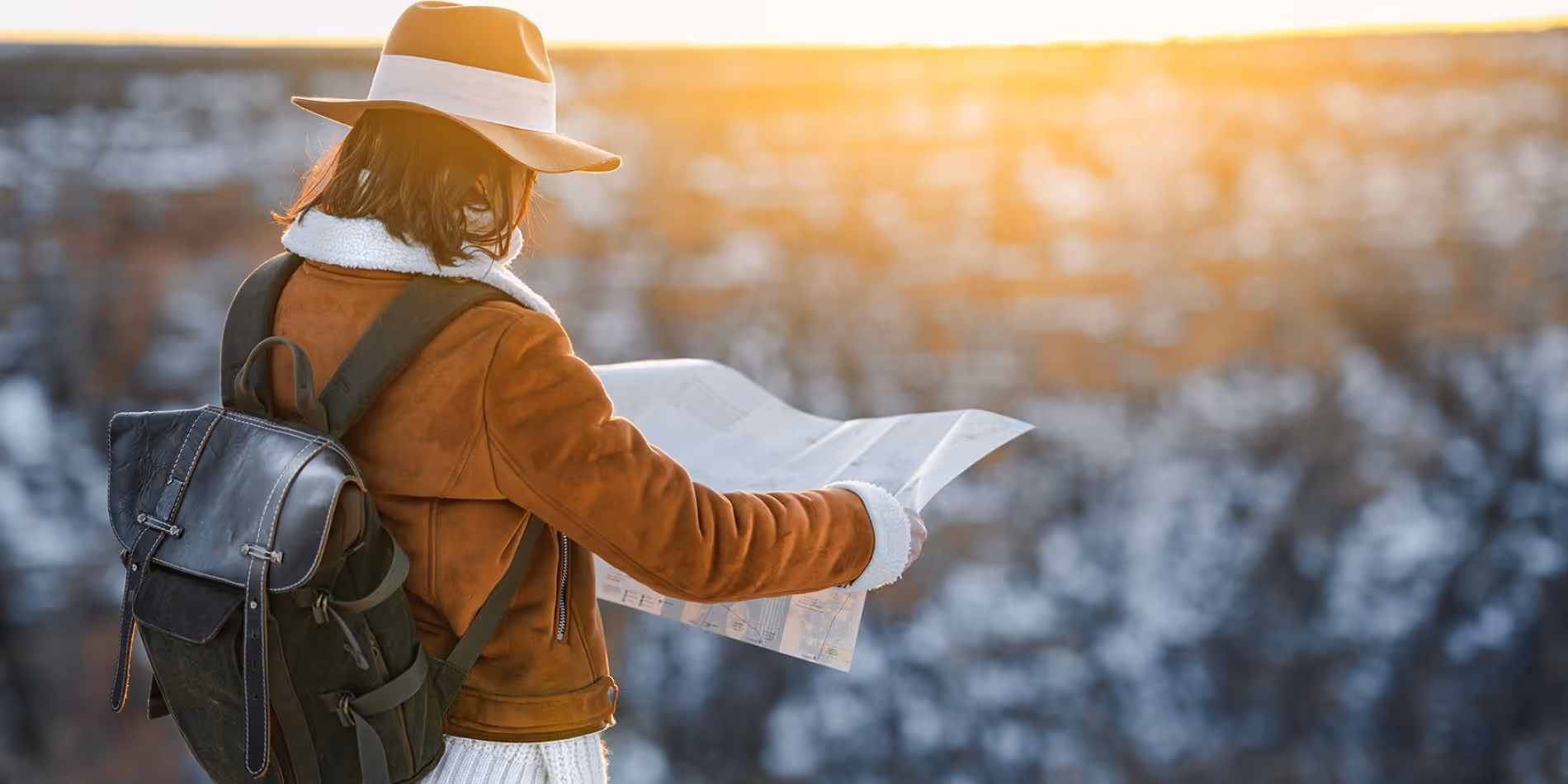 Person wearing a tan hat and brown jacket with a backpack looks at a map with a snowy landscape and sunset in the background.