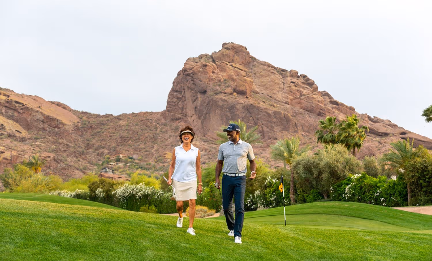 A man and woman walking on a green golf course with rocky mountains and palm trees in the background.