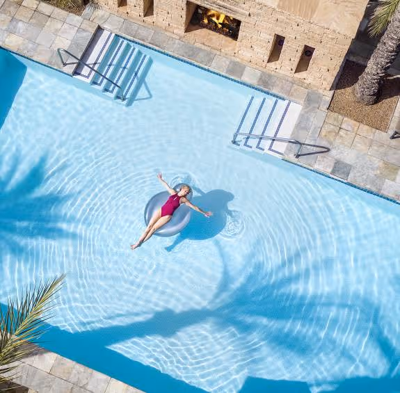 Woman in a pink swimsuit relaxing on an inflatable ring in a clear blue swimming pool with palm tree shadows and a stone fireplace nearby.