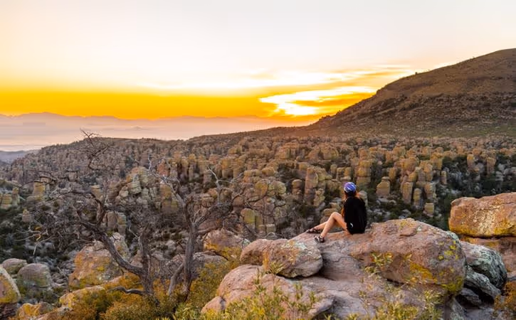 Person sitting on rocky terrain overlooking a rugged landscape at sunset with vibrant orange sky.
