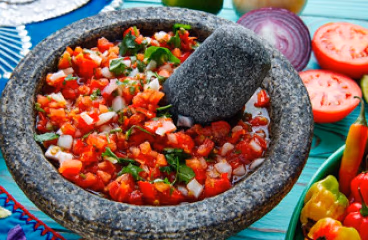 Stone molcajete filled with freshly made salsa containing diced tomatoes, onions, and cilantro with a stone pestle inside.