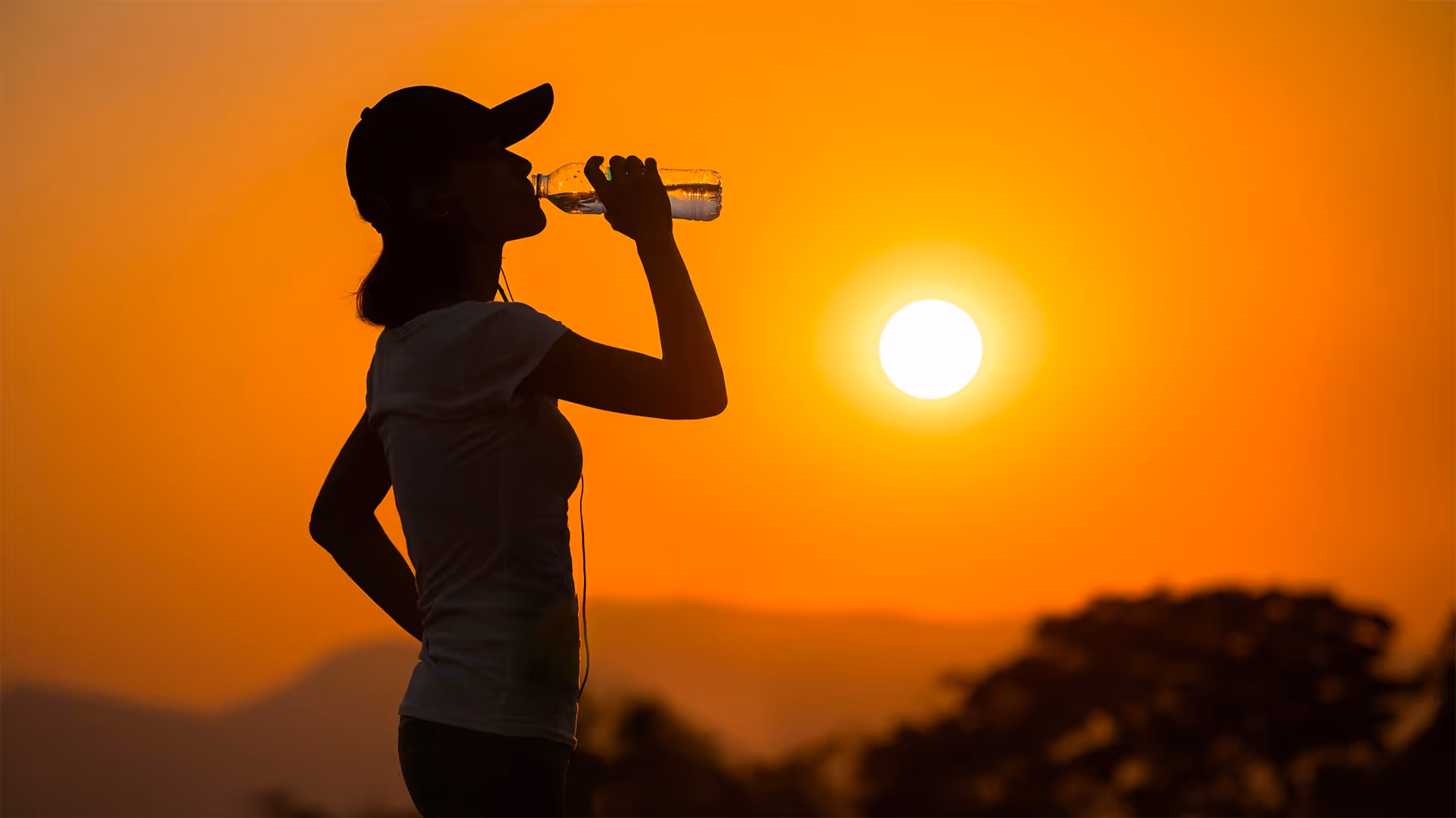 Silhouette of a person drinking water from a bottle at sunset with an orange sky and distant hills.