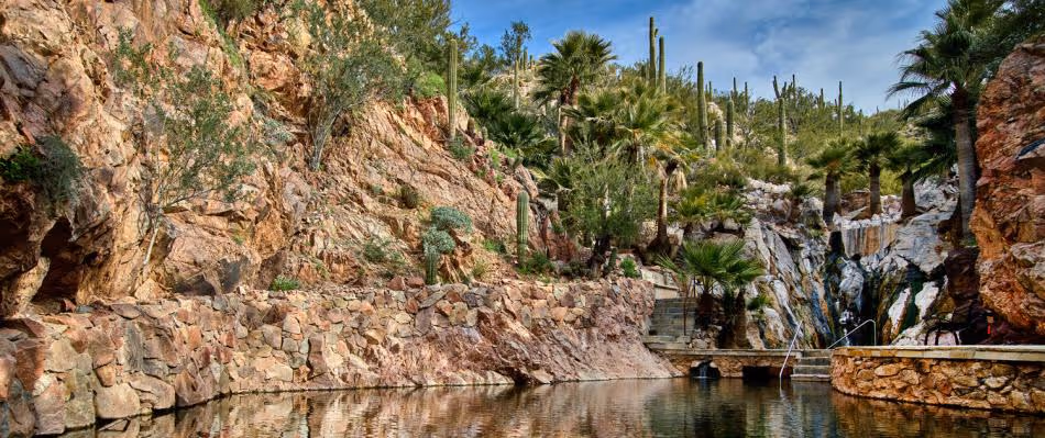 Natural rock pool surrounded by rugged cliffs, desert vegetation, and palm trees under a partly cloudy sky.