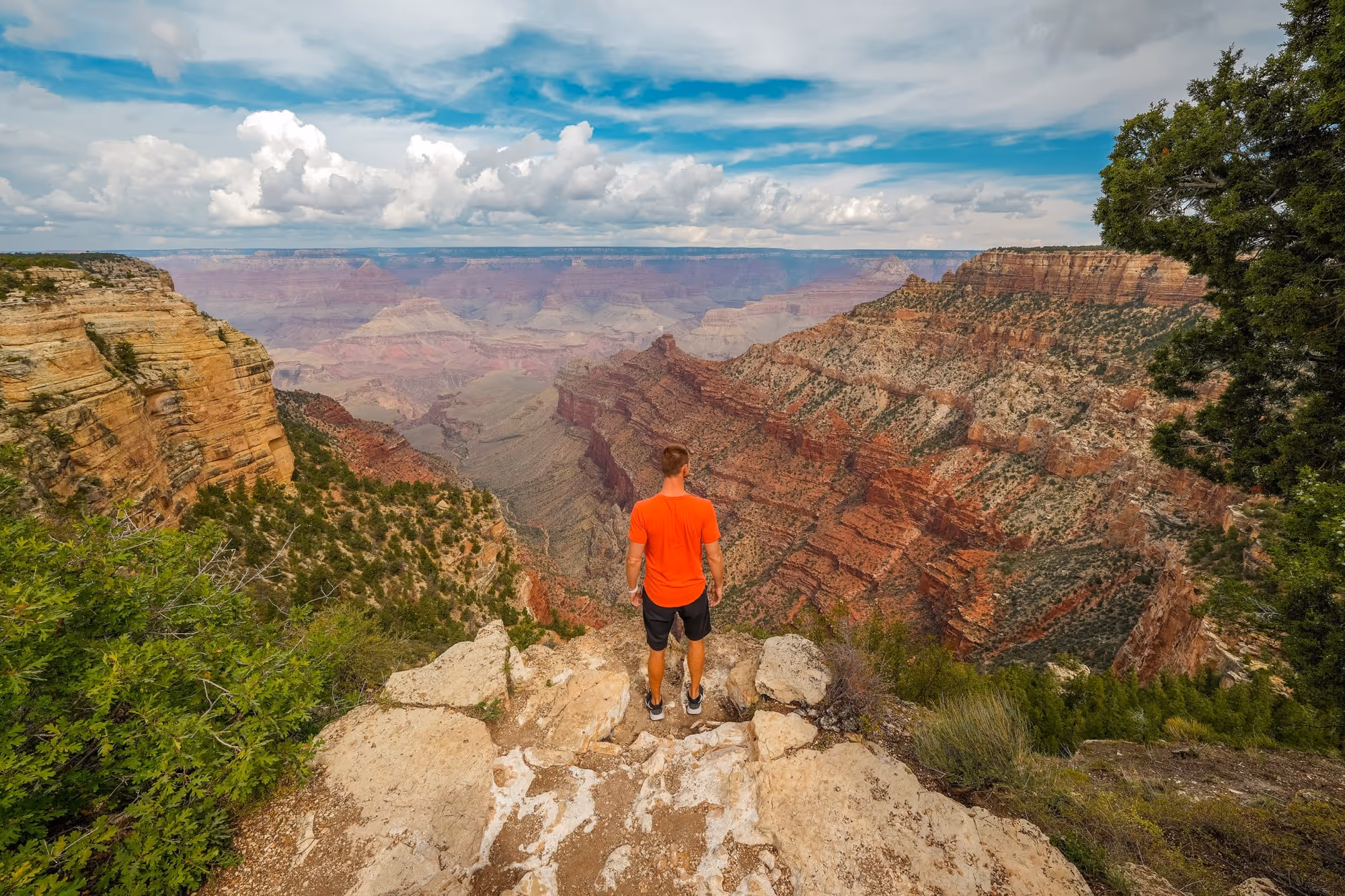 Man in orange shirt standing on rocky cliff overlooking the expansive Grand Canyon under a partly cloudy sky.