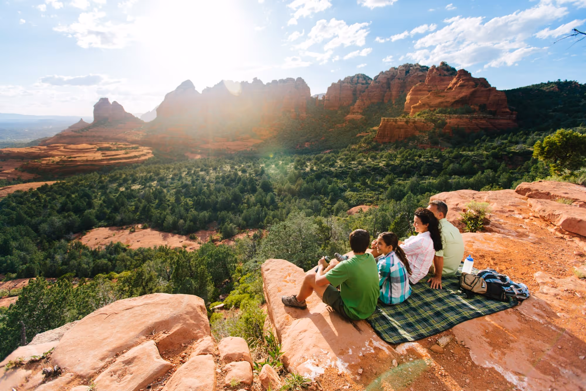Family of four sitting on a blanket on a red rock cliff overlooking a green forest and red rock formations with the sun shining in a partly cloudy sky.