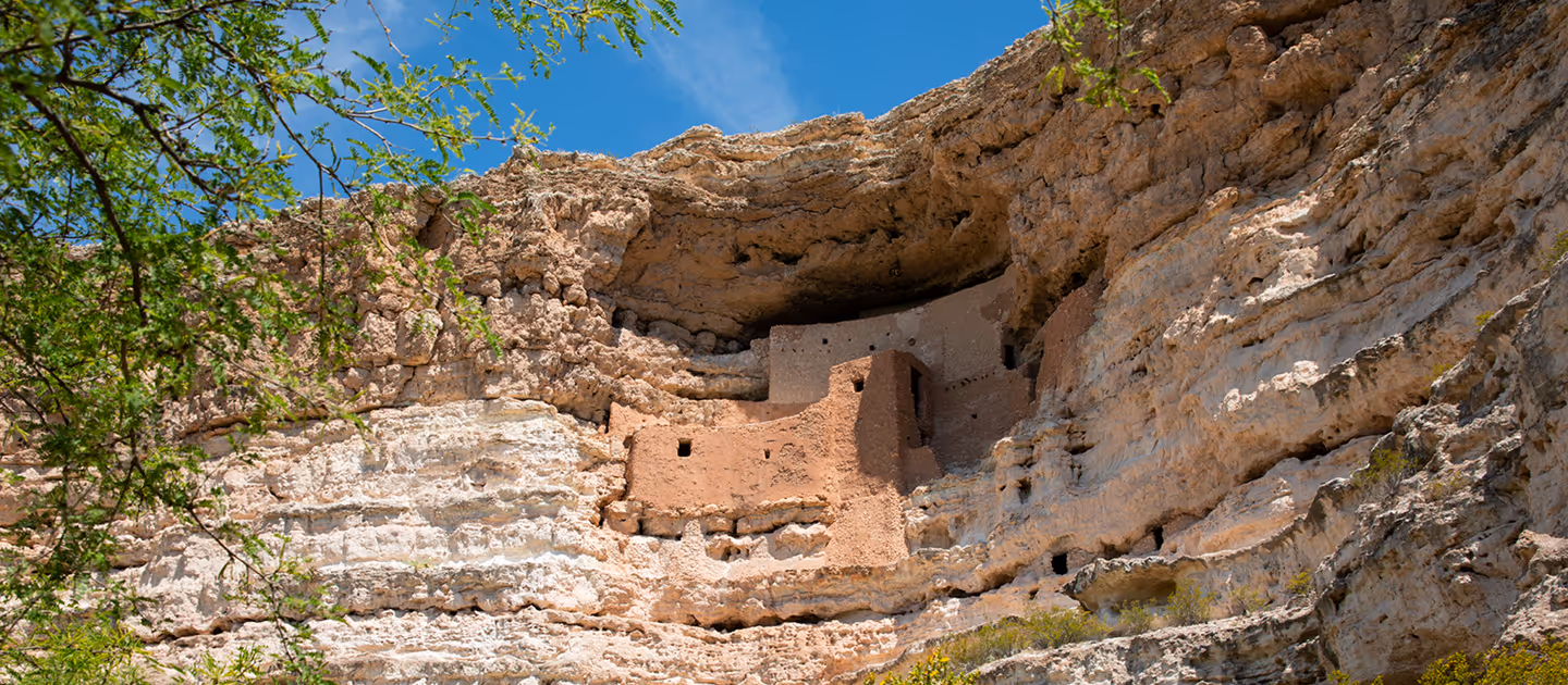 Ancient cliff dwellings built into a rocky overhang with green foliage in the foreground under a blue sky.