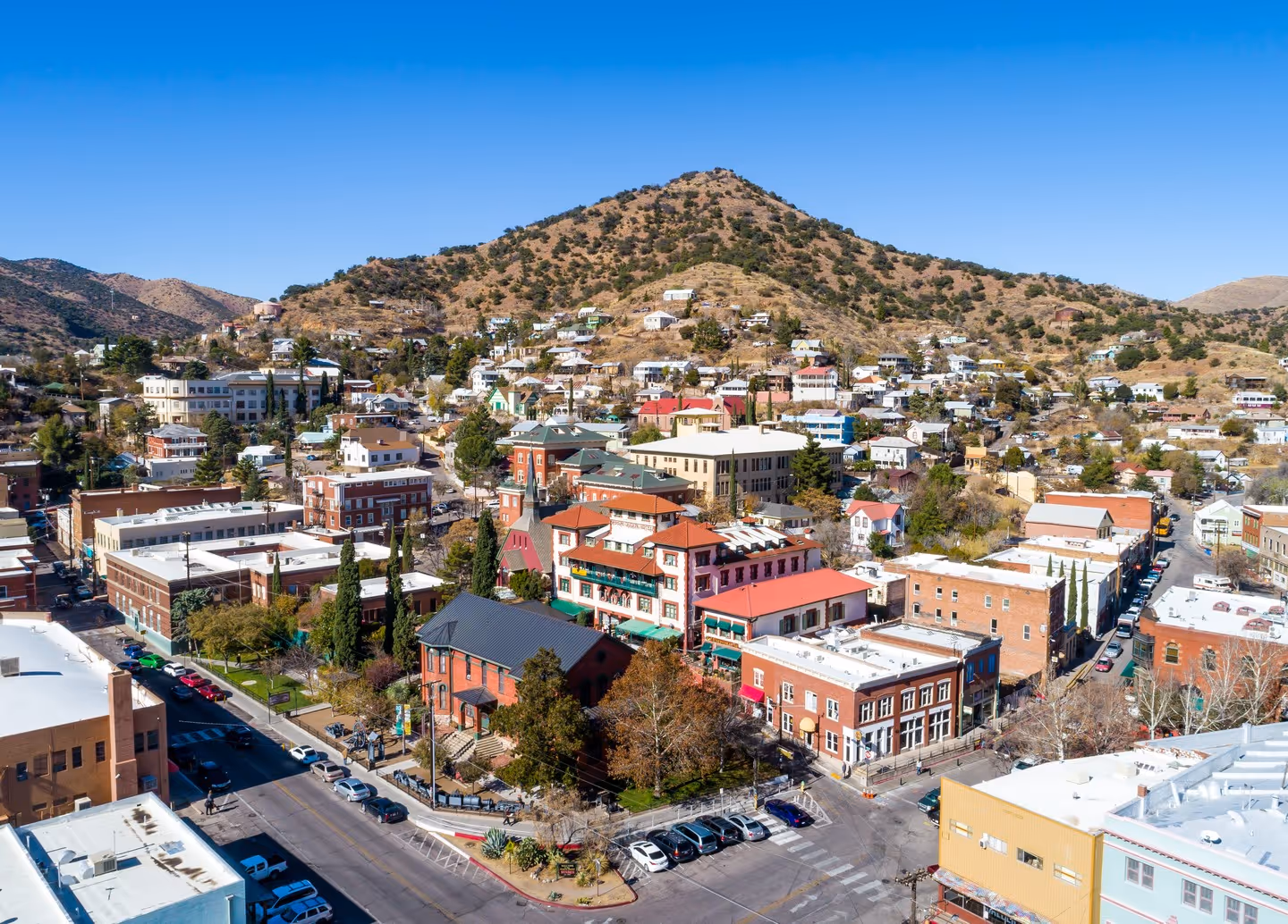 Aerial view of Bisbee, Arizona with brick buildings, parked cars, and a hillside dotted with houses under a clear blue sky.
