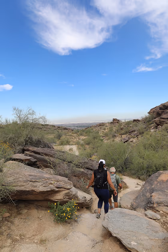 Two hikers walking down a rocky desert trail surrounded by scrub and bushes under a partly cloudy blue sky.