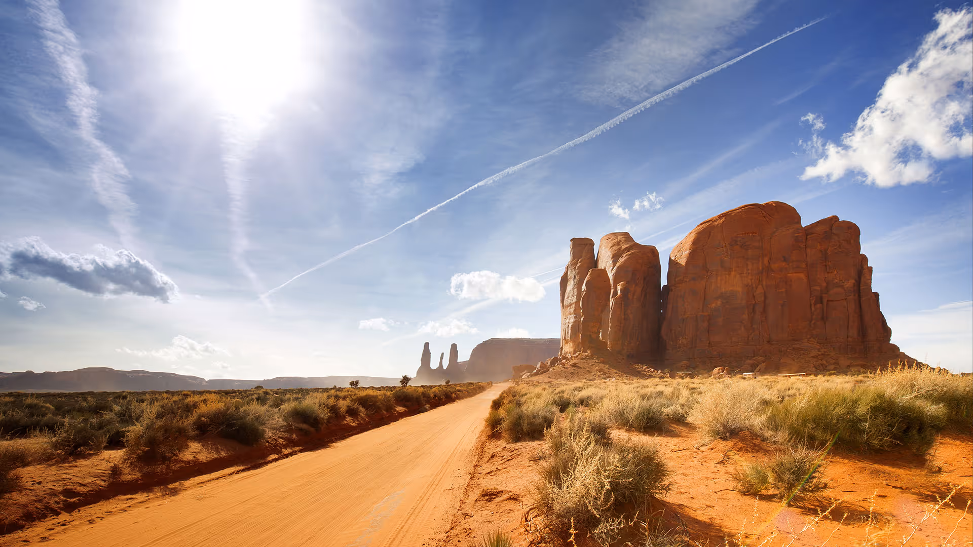Sunlit red rock formation beside a dirt road under a blue sky with scattered clouds in a desert landscape.