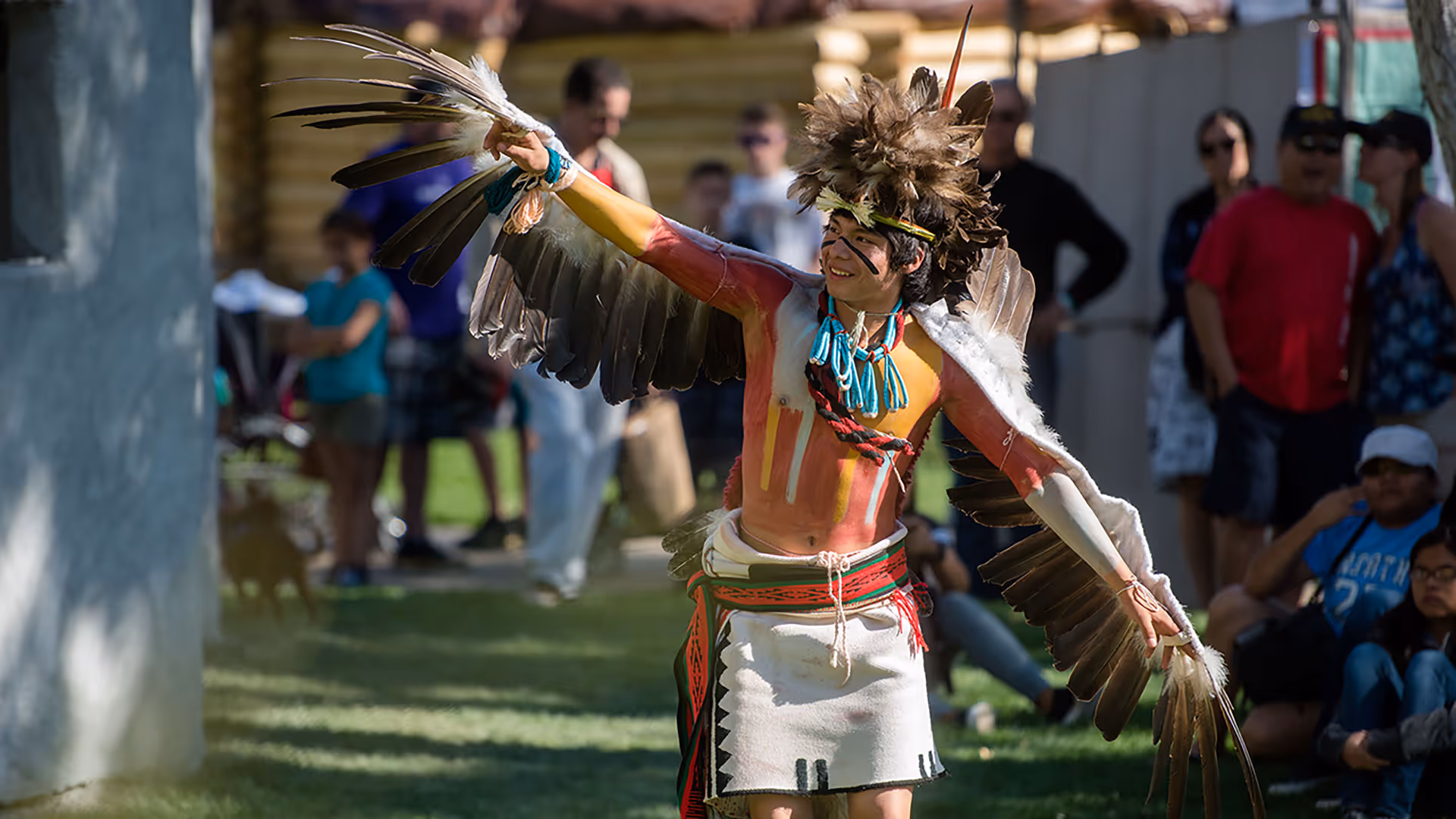 Young man performing a traditional Indigenous dance wearing feathered headdress, winged arm decorations, and face paint.