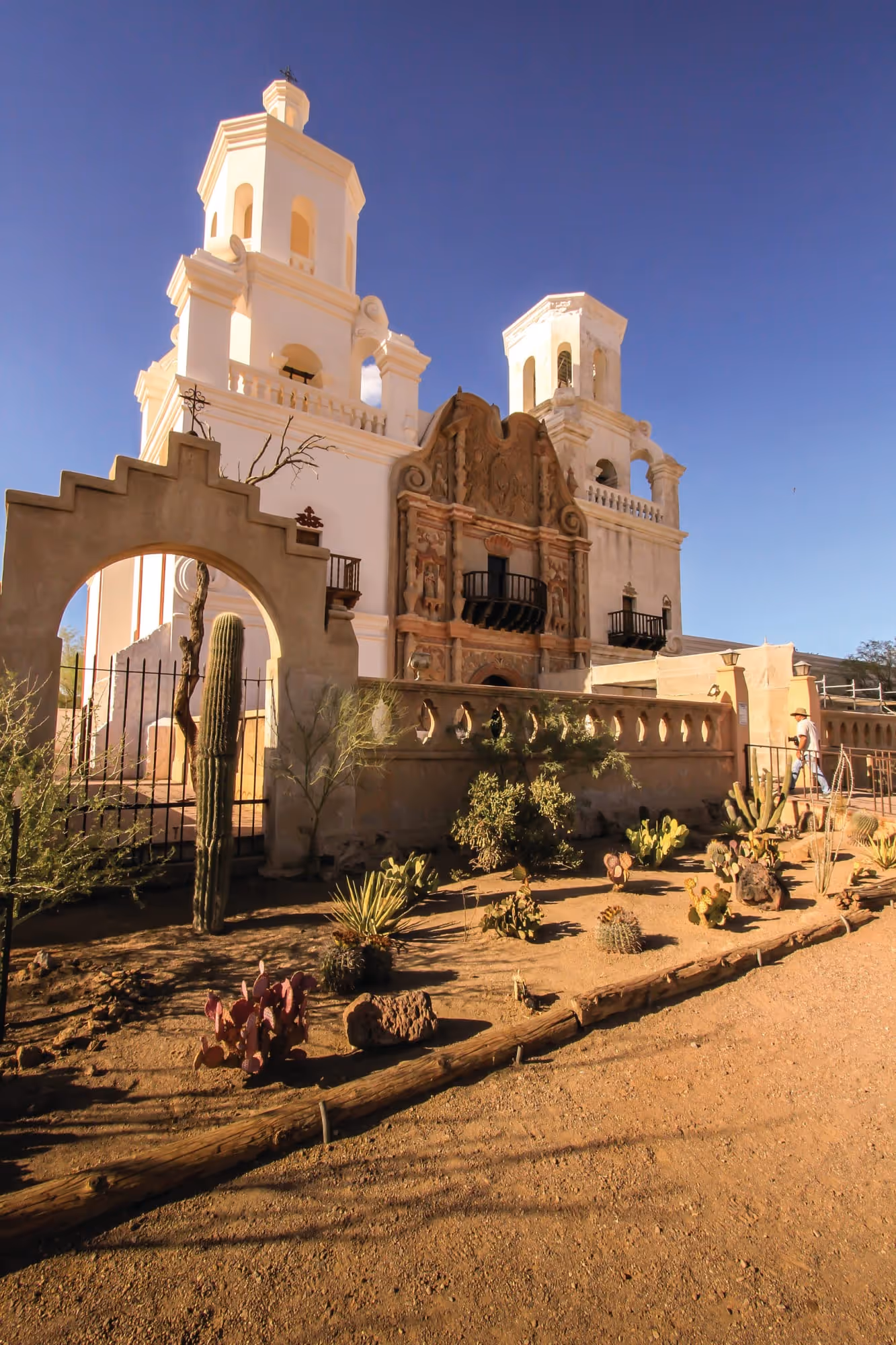 View of historic San Xavier del Bac Mission with white towers and desert plants in front under clear blue sky.