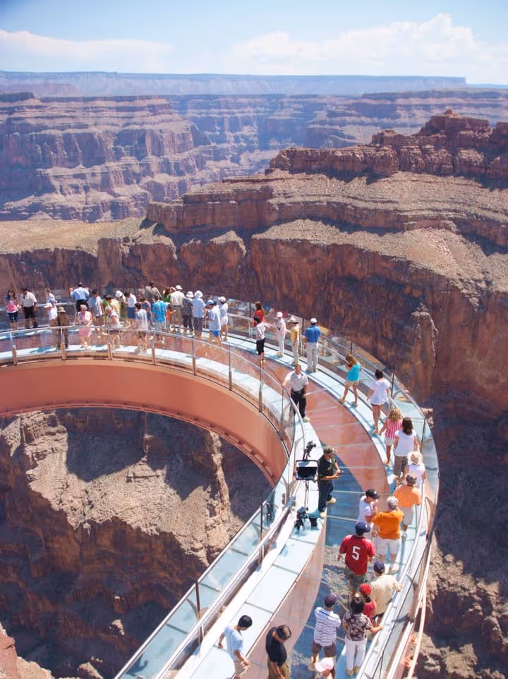 People walking on the Grand Canyon Skywalk, a curved glass bridge extending over a deep canyon with layered rocky cliffs under a clear sky.