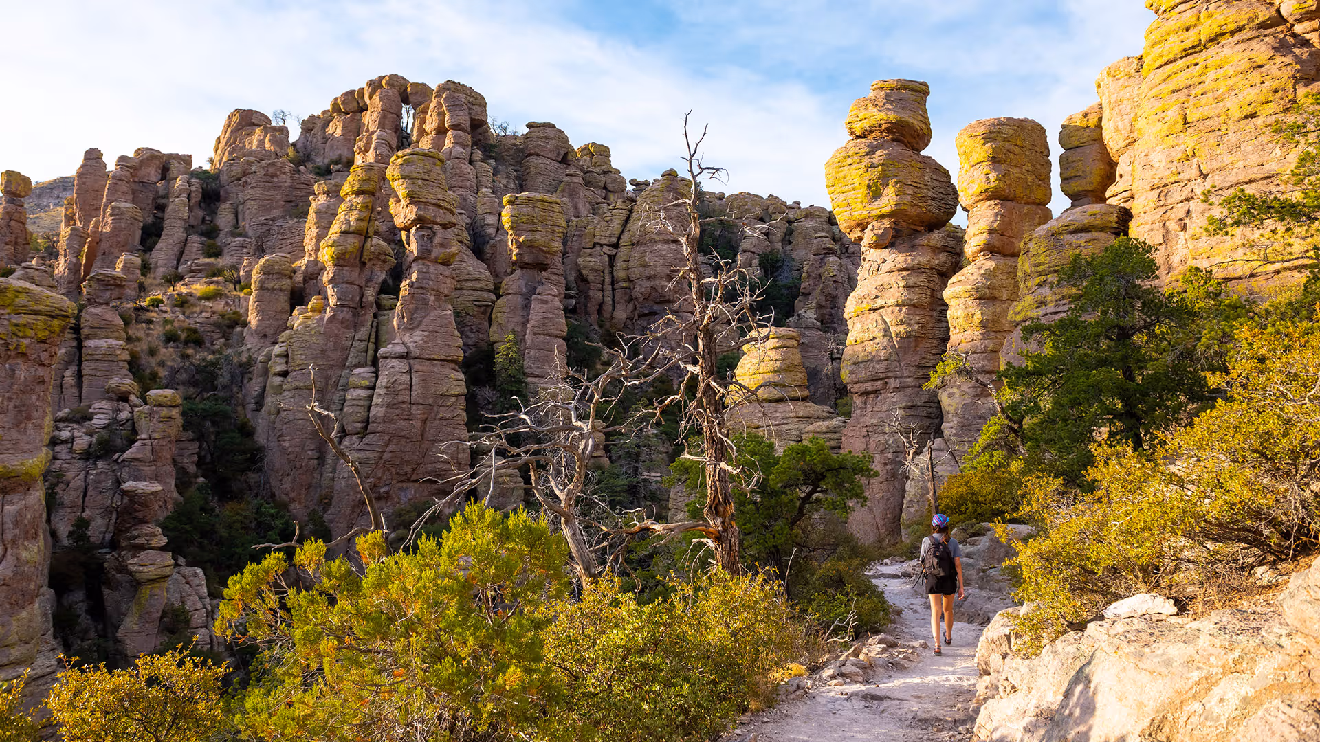 Hiker walking on a trail surrounded by tall, uniquely shaped rock formations and vegetation under a partly cloudy sky.