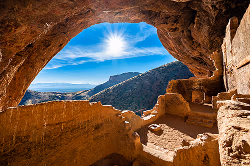 Sunlit ancient cliff dwellings inside a rocky cave overlooking a mountainous landscape under a bright blue sky.