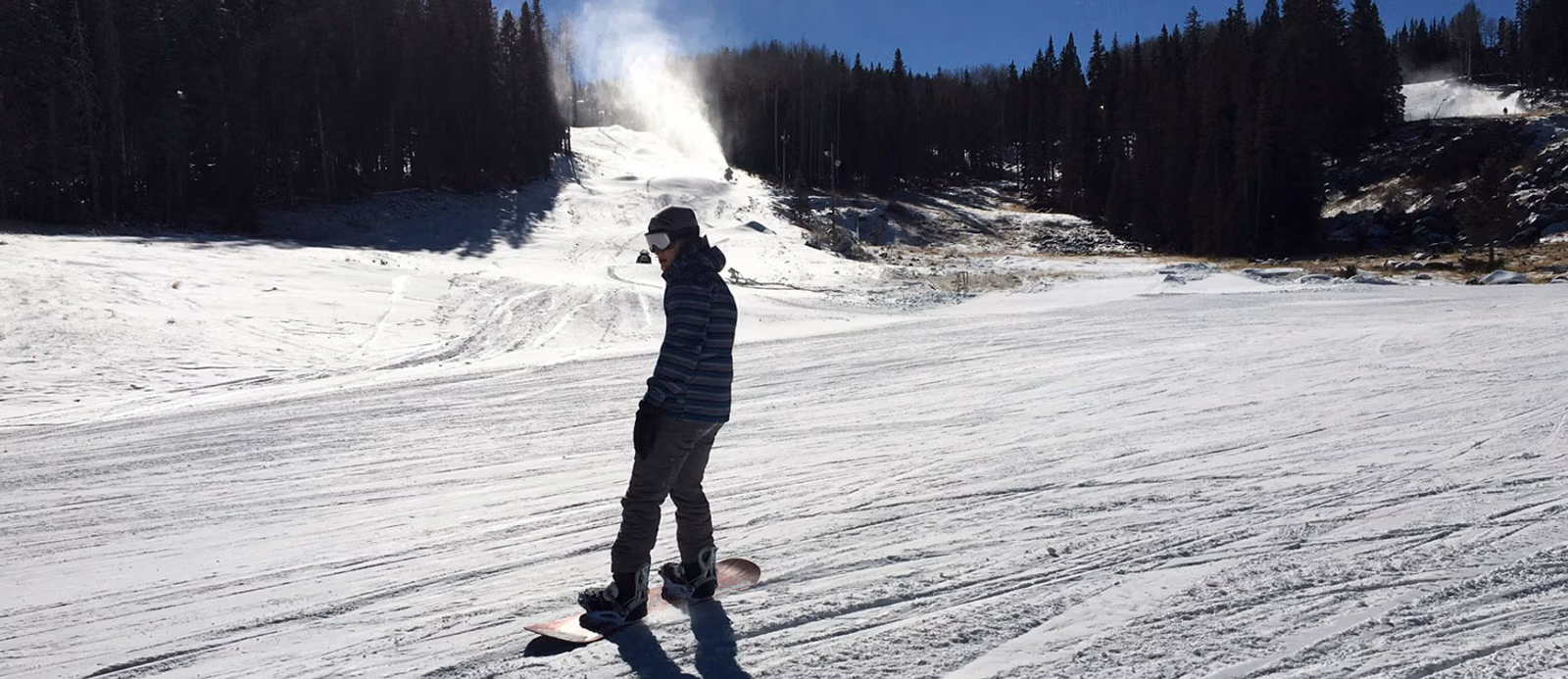 Person snowboarding on a snowy slope at a ski resort with snow machines and pine trees in the background.