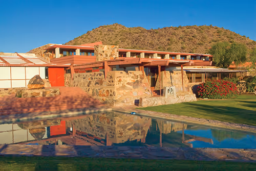 Stone and wood building with red accents reflected in a pool, set against a clear blue sky and desert hill.