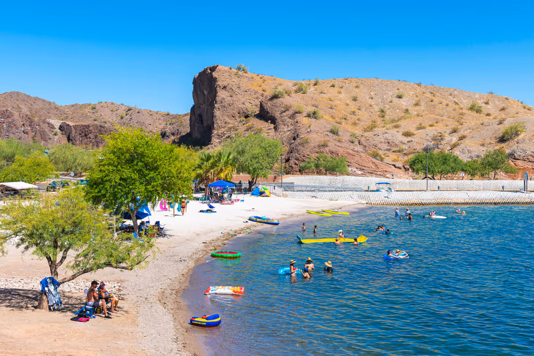 People swimming and relaxing on a sunny sandy beach by a lake with rocky hills in the background.