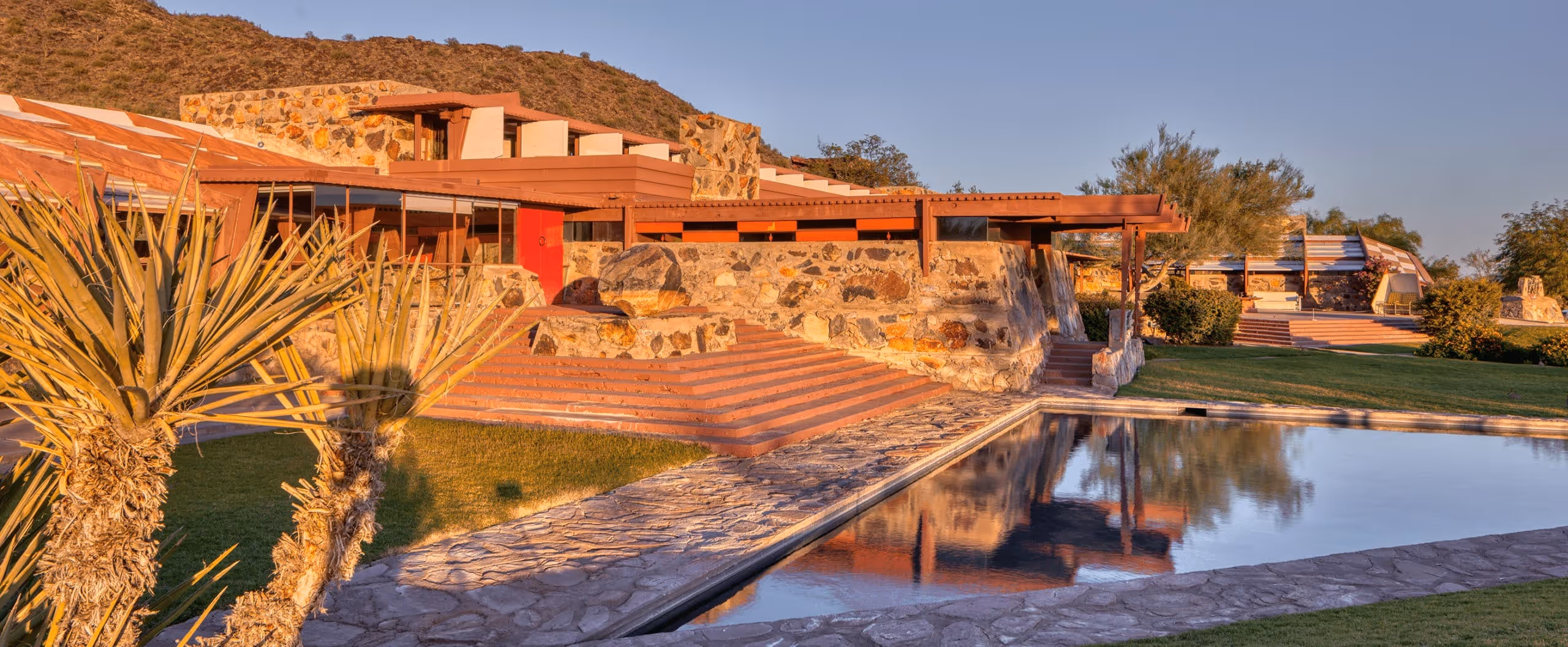Stone and wood building with red accents beside a reflective swimming pool, surrounded by desert plants and hills under clear sky.
