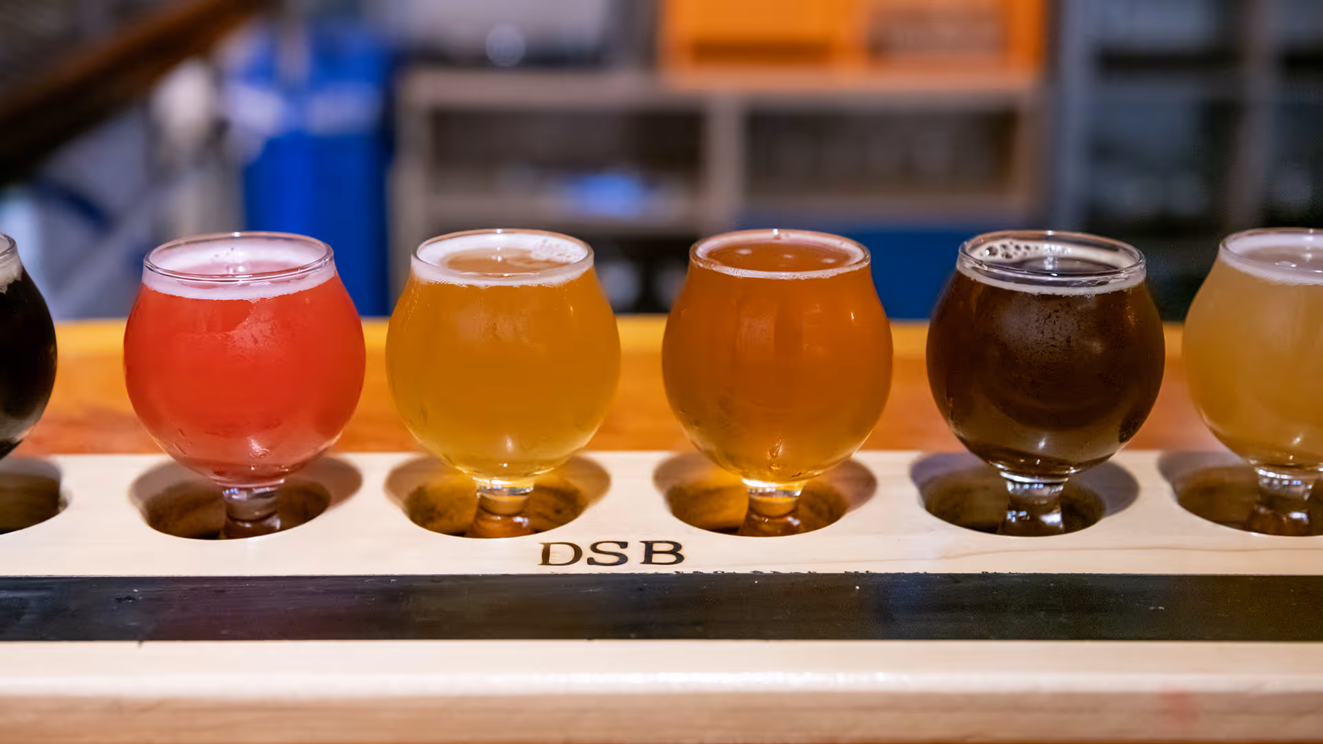 A flight of five craft beers in small round glasses on a wooden holder engraved with 'DSB'.
