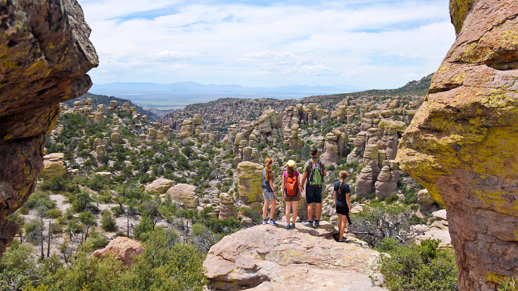 Four hikers standing on a large rock overlooking a vast landscape of rock formations and sparse greenery under a partly cloudy sky.