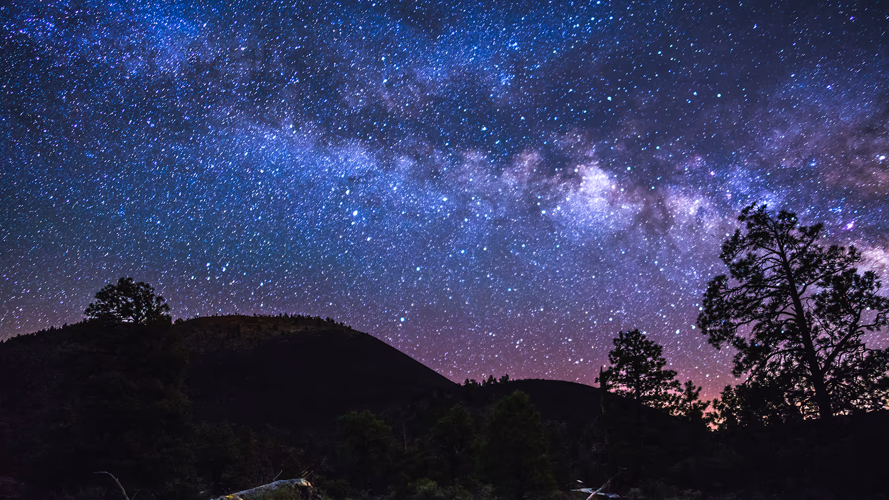 Night sky filled with stars and the Milky Way galaxy over dark mountain silhouettes and trees.