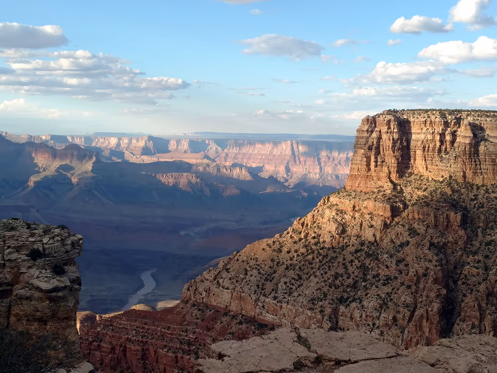 View of the Grand Canyon with layered rock formations, deep shadows, and a river winding through the canyon under a partly cloudy sky.