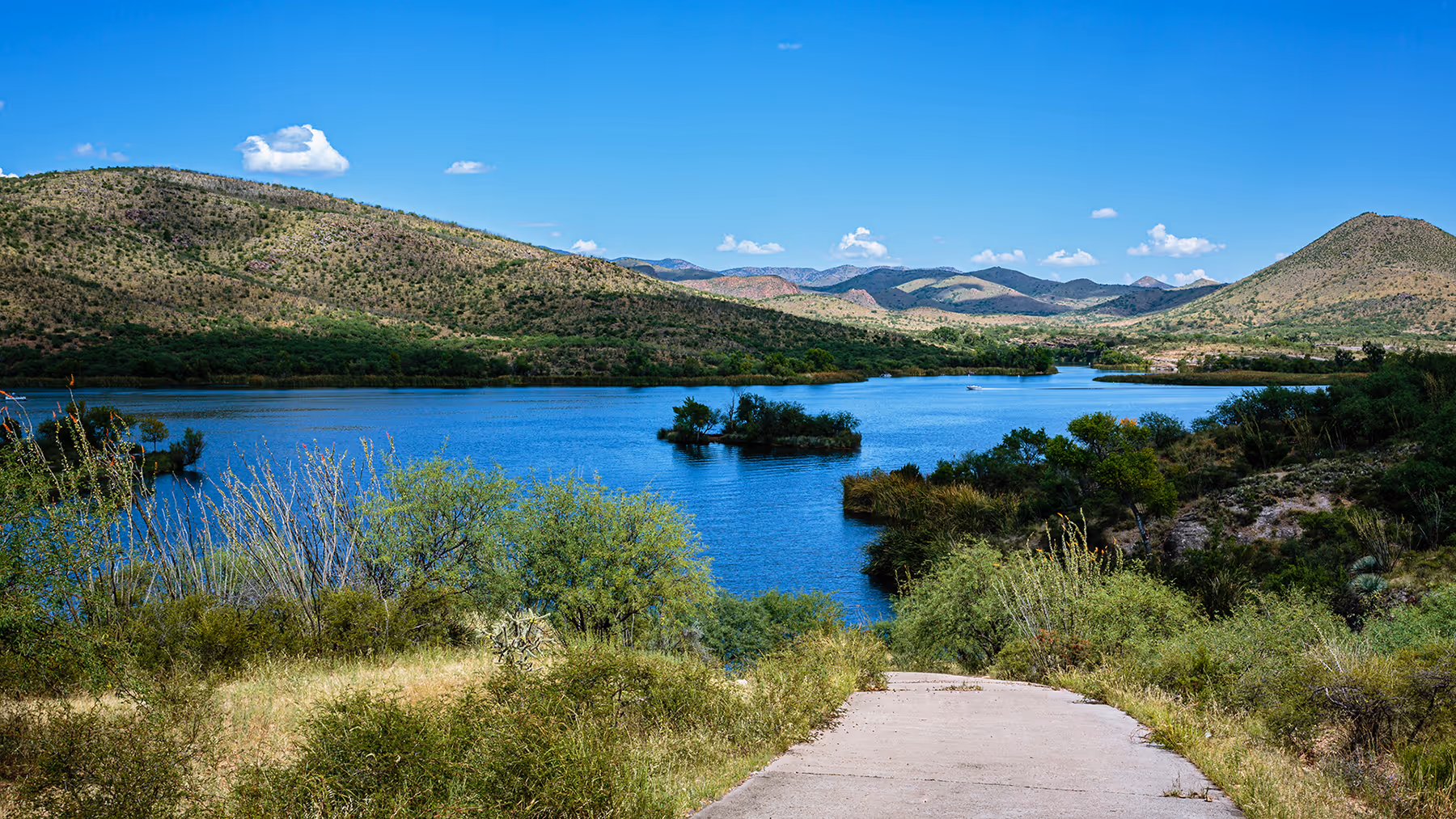 Path leading to a blue lake surrounded by green bushes and hills under a clear blue sky.