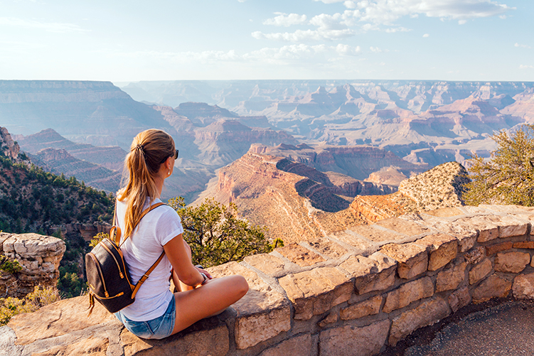 Woman with backpack sitting on stone wall, overlooking vast Grand Canyon landscape under blue sky.