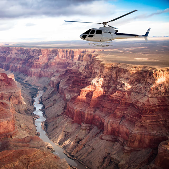 White helicopter flying over the Grand Canyon with a river winding through rocky cliffs below.