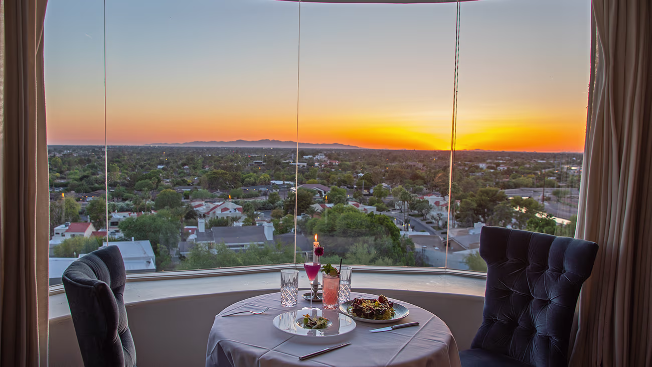 A close-up of a dinner table for two set next to a curved window, overlooking the city of Phoenix and distant mountains during a vibrant sunset. The table has a white cloth, plates of food, cocktails and a candle. Two tufted velvet chairs face the golden-orange sky.
