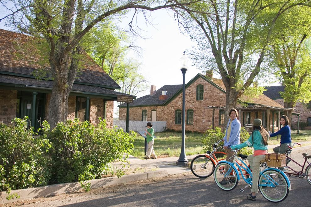 Three people on bicycles smiling near historic stone buildings with tall trees and a child reading a sign.