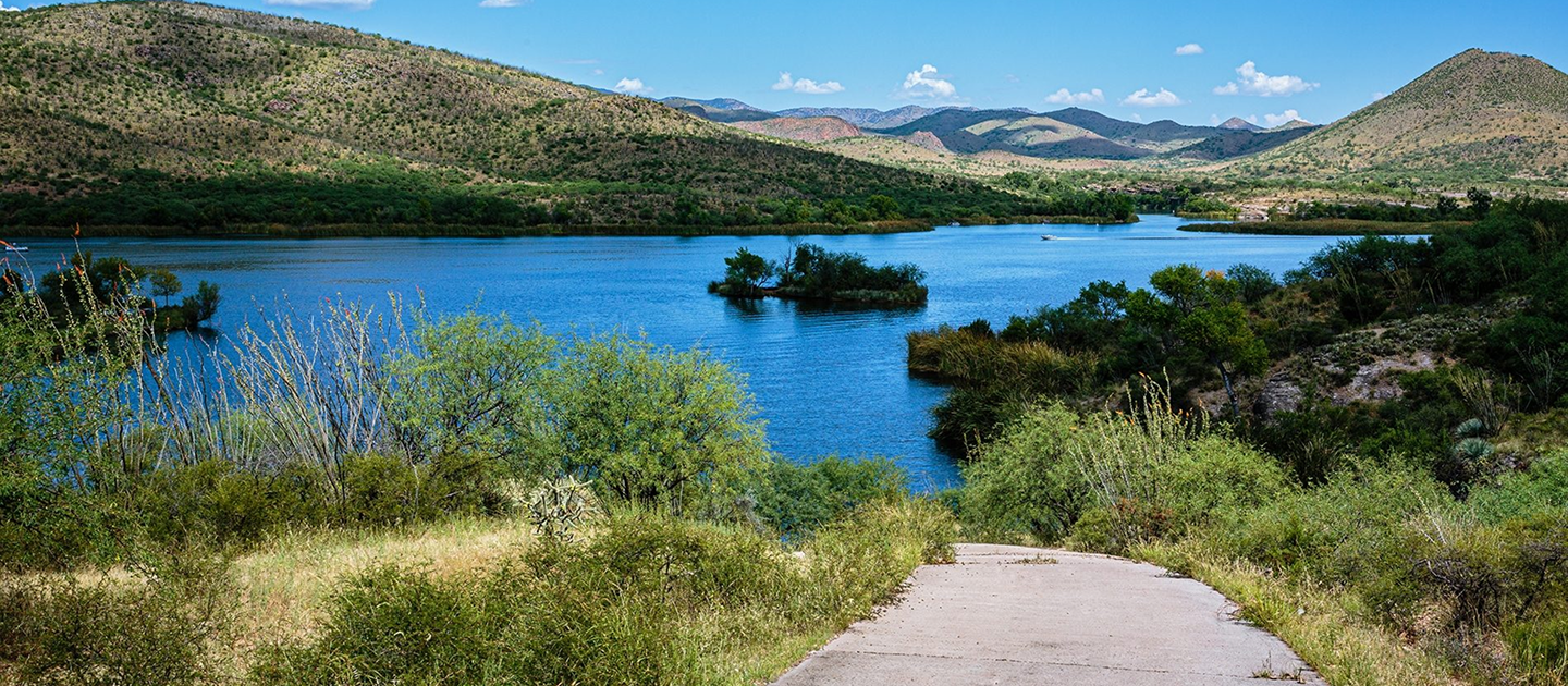 Path leading to a bright blue lake with small islands and surrounding green hills under a clear sky.