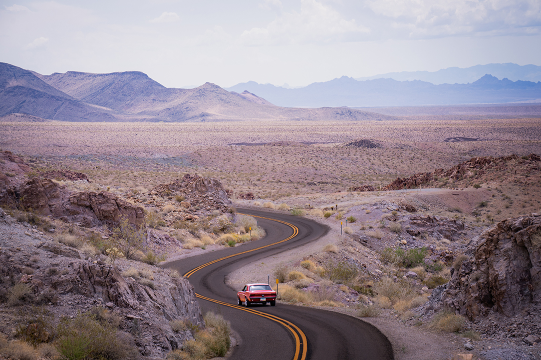 Red car driving on winding road through rocky desert landscape with distant mountains under cloudy sky.