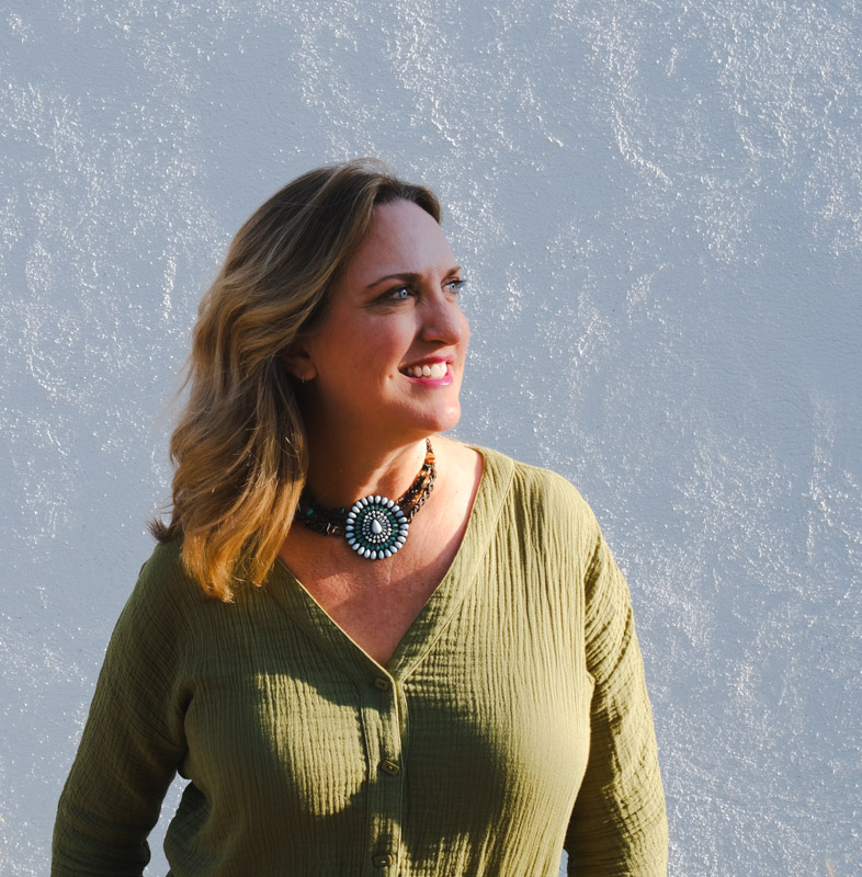 Woman with light brown hair wearing an olive green blouse and decorative necklace, smiling looking right.