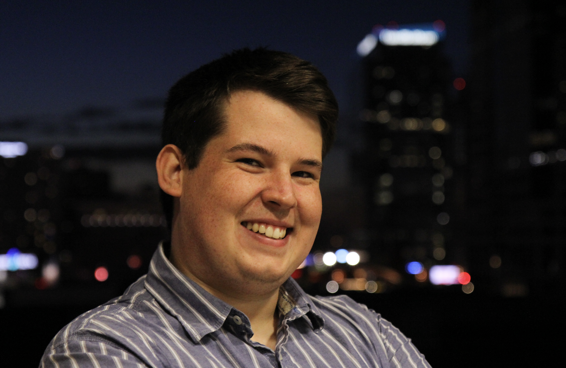 Smiling young man in a striped shirt with arms crossed, nighttime city lights blurred behind him.