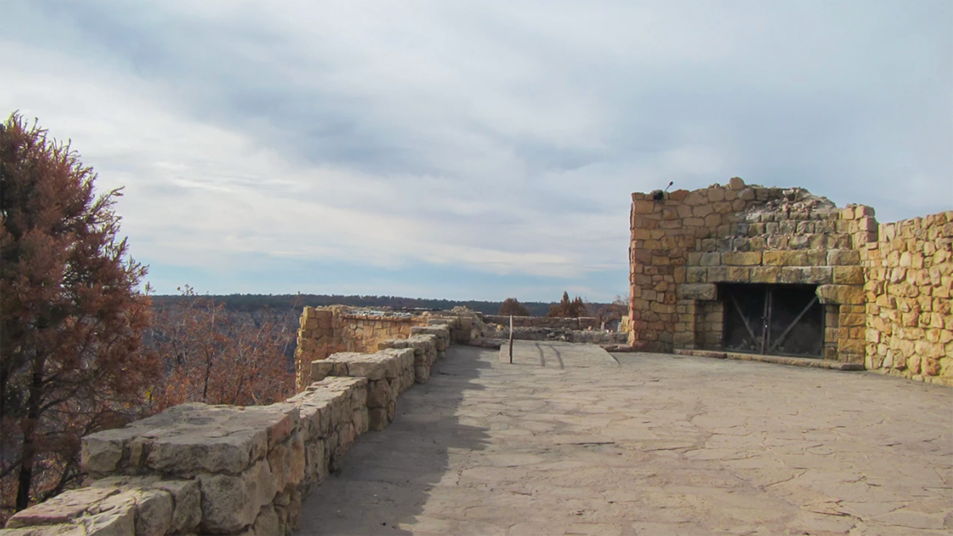Stone terrace with low walls and large stone fireplace overlooking forested canyon under cloudy sky.
