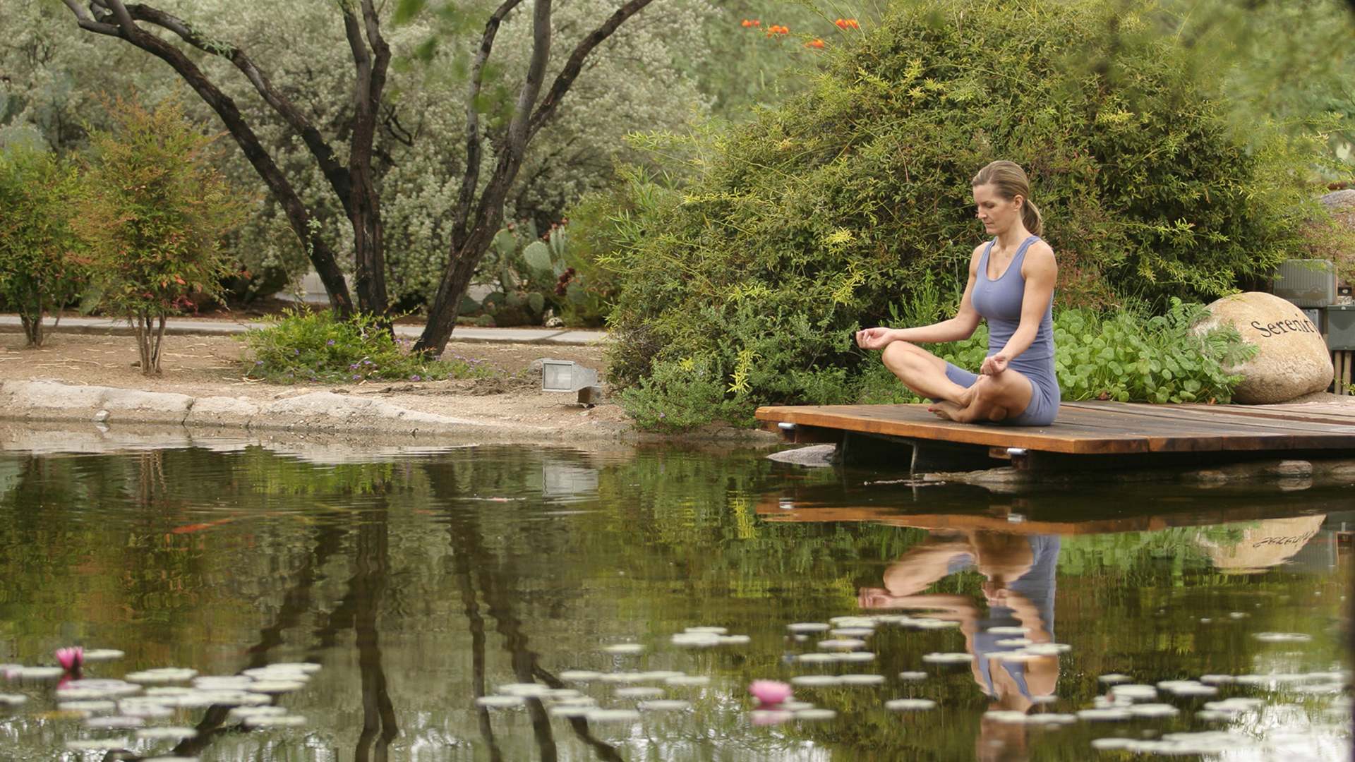 Woman meditating in lotus pose on wooden platform by pond with lily pads and surrounding greenery.