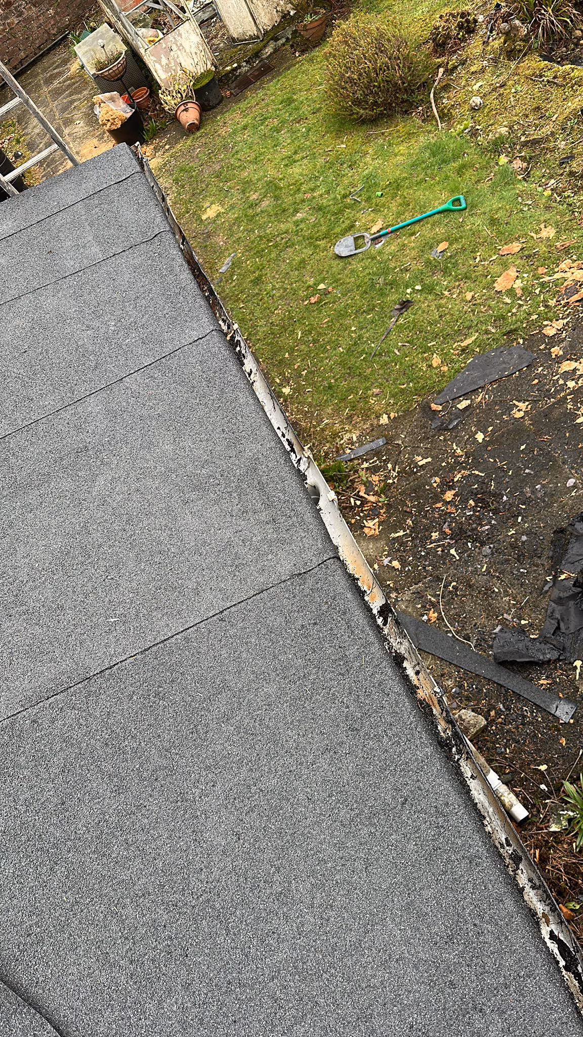 Overhead view of a flat gray rooftop next to a garden with green grass, a green-handled shovel, plant pots, and various gardening debris.